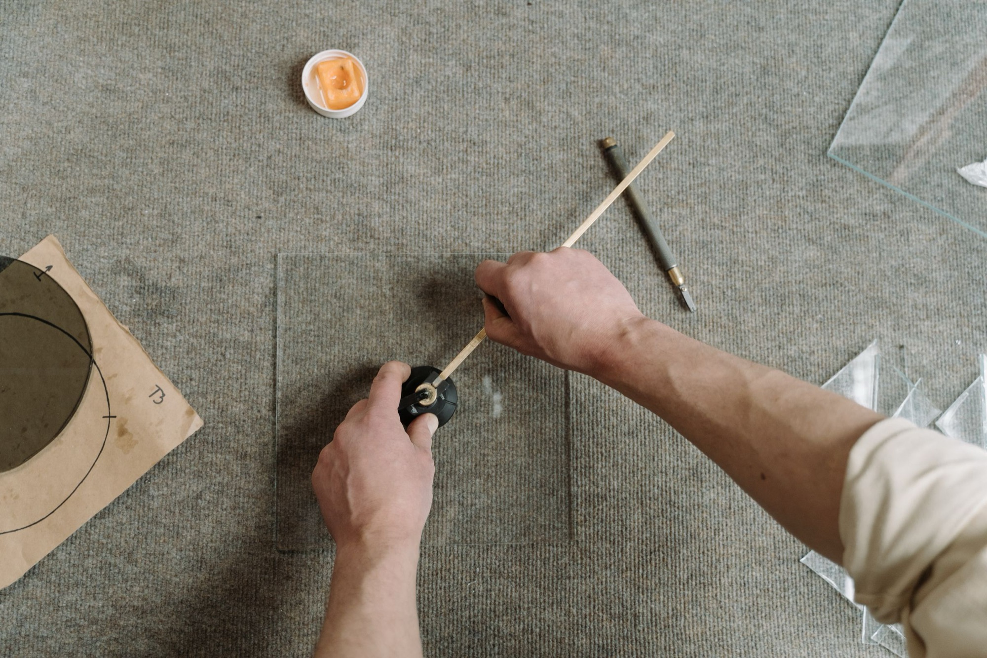 Hands scoring and measuring a sheet of glass on a workshop table. Illustrative photograph.