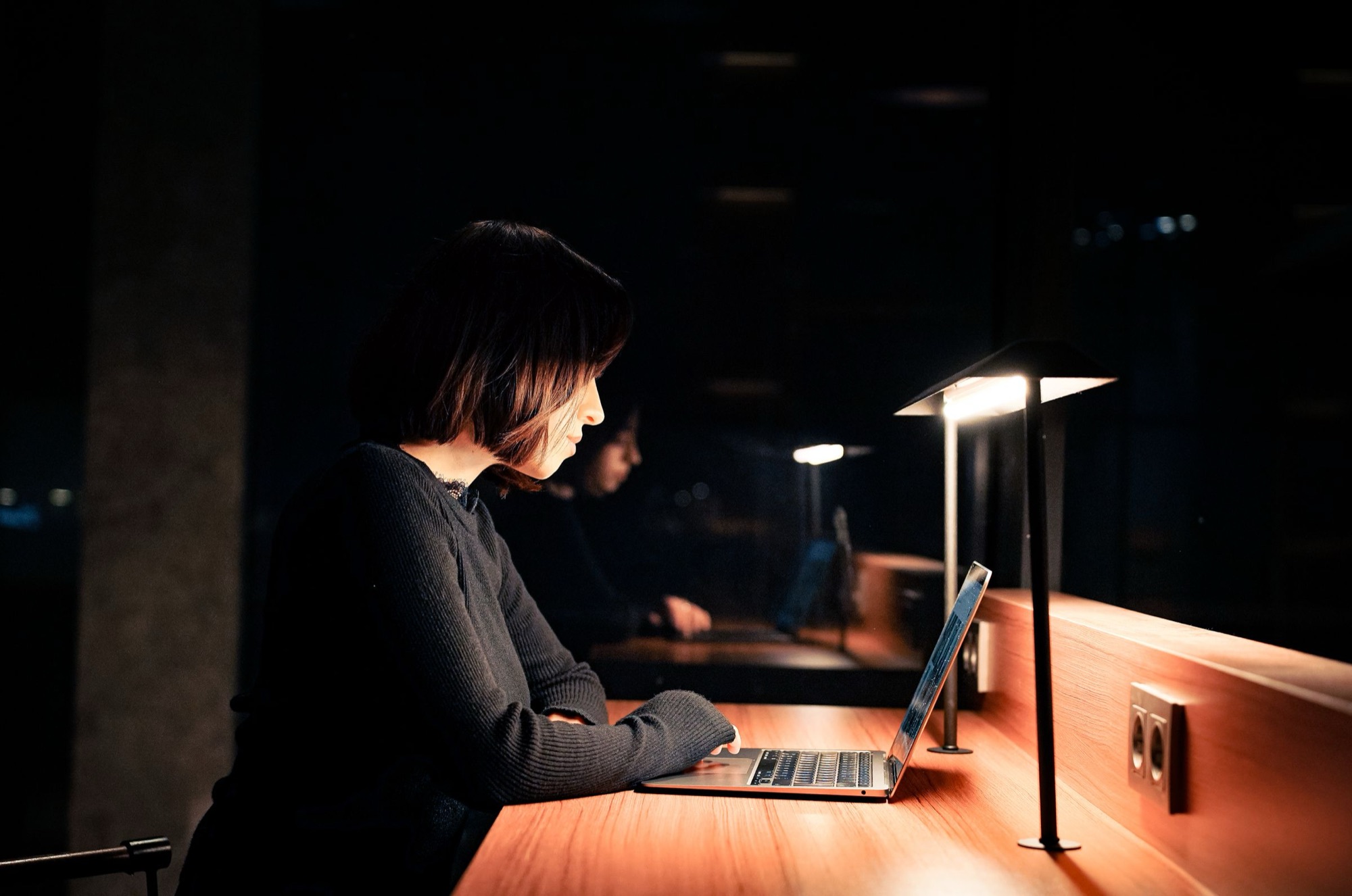 Someone working at a laptop under a warm desk lamp late in the evening. Illustrative photograph.