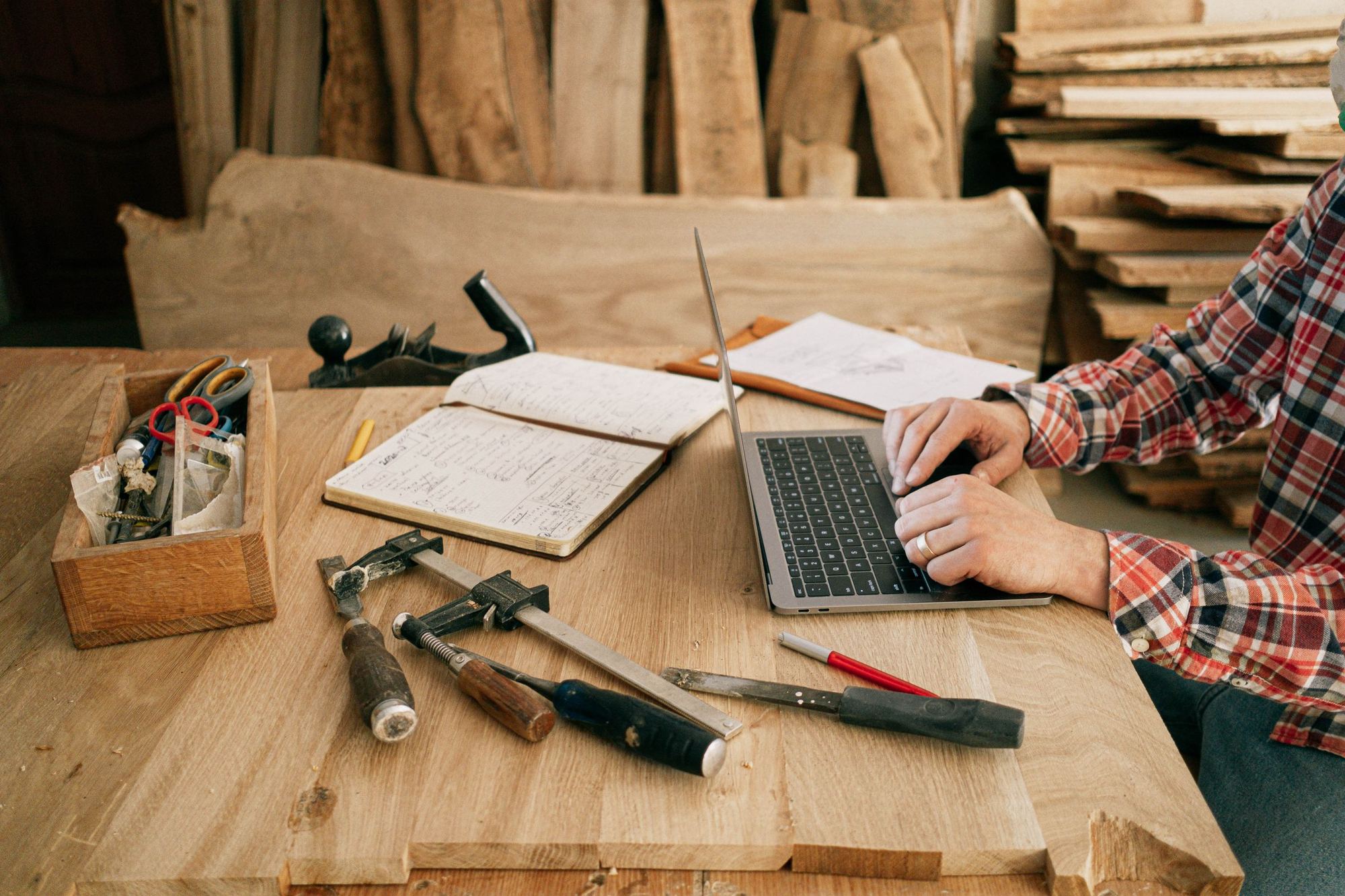 A woodworker at a laptop on a workbench with tools and a notebook.
